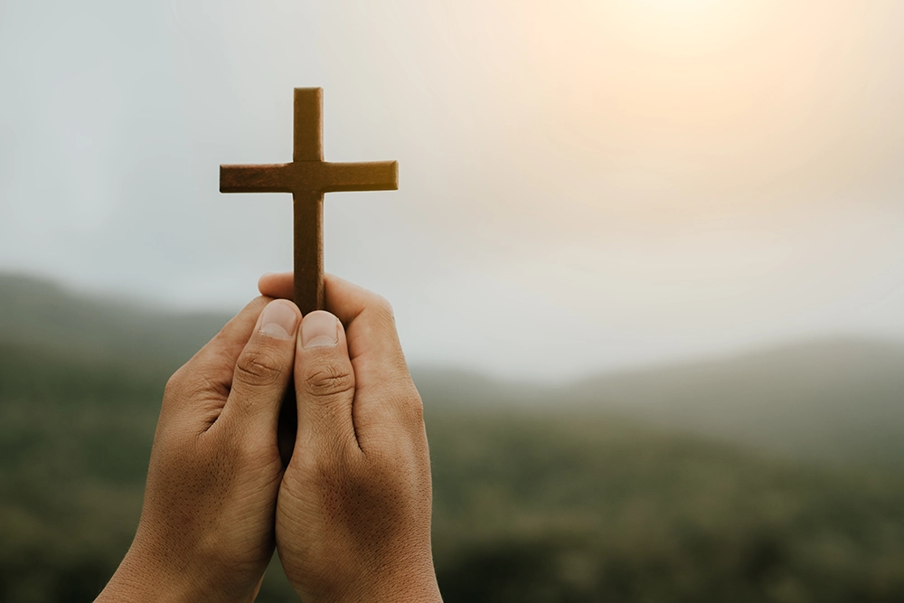 man-praying-with-cross-in-nature-sunrise-backgroun-2023-11-27-04-56-45-utc hands holding a wooden cross outside