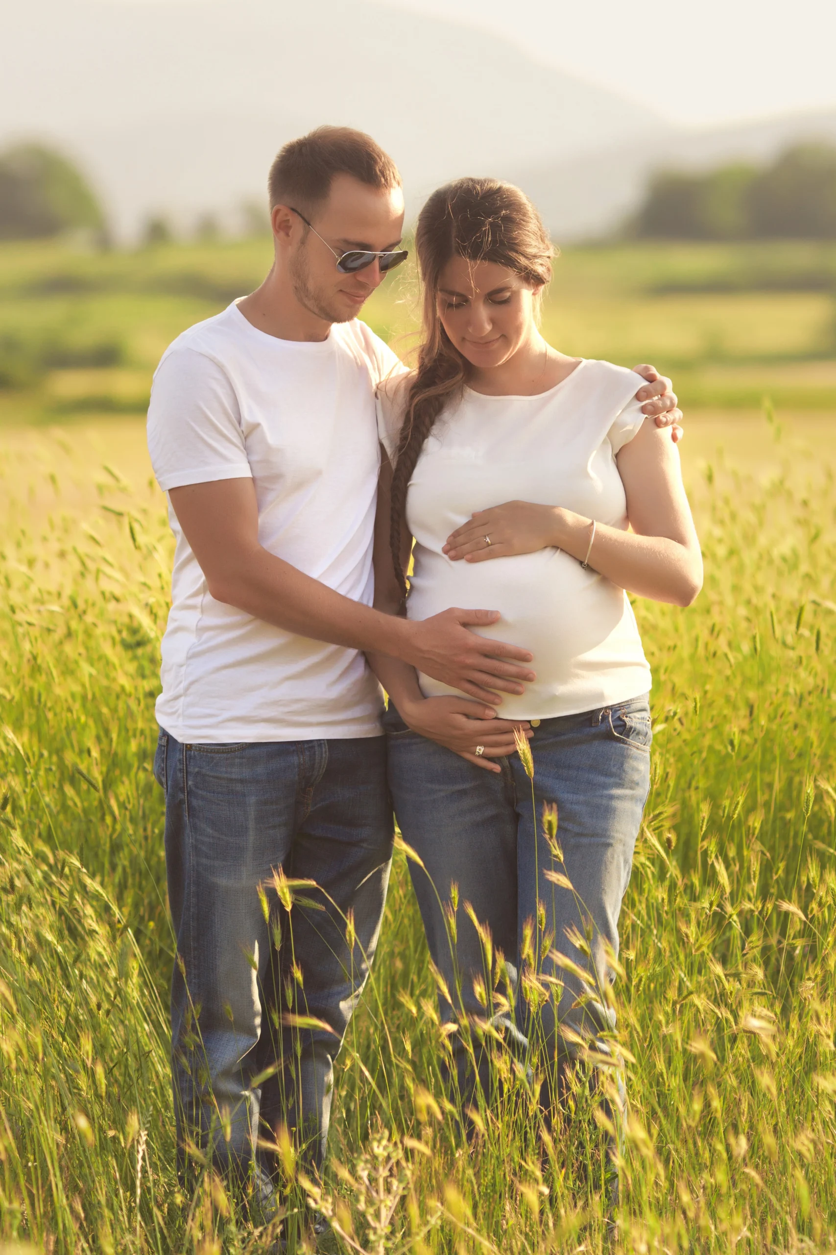 couple standing in a field embraced with hands on woman's pregnant belly.