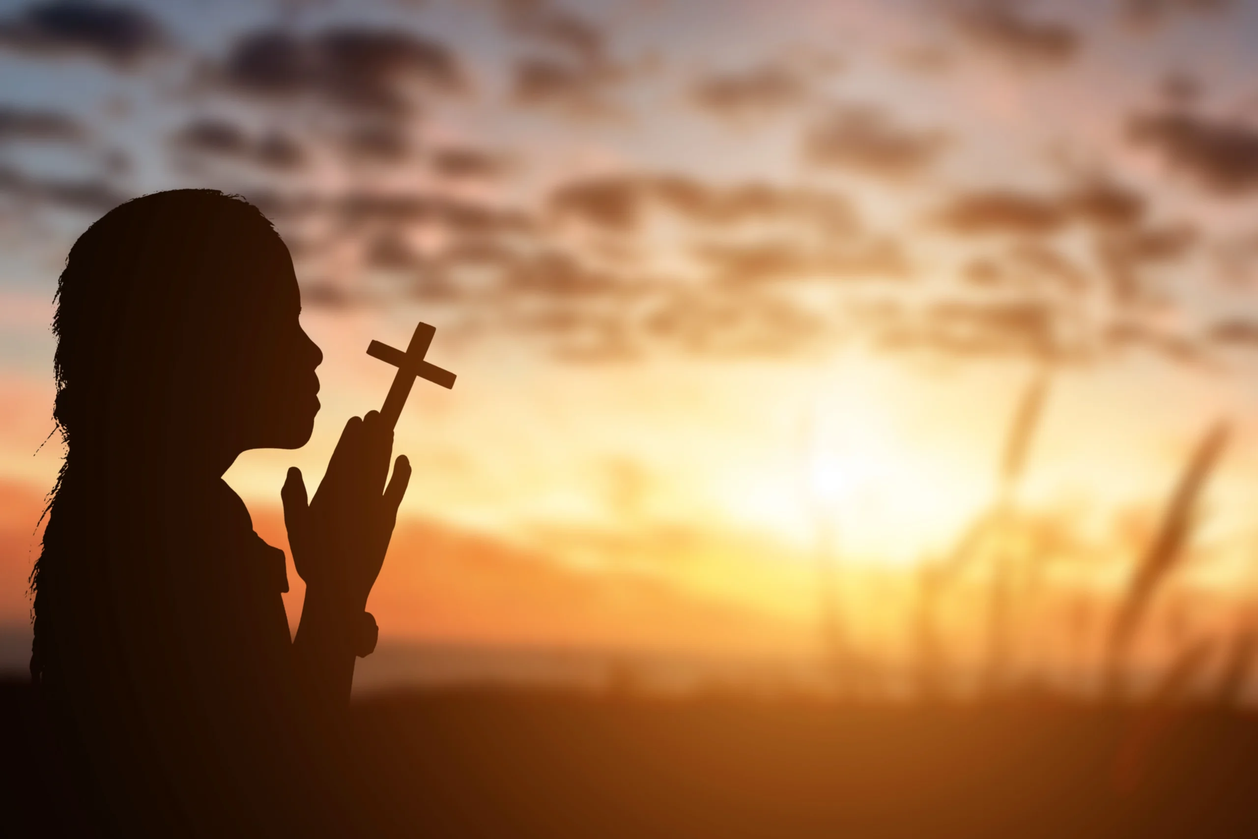 little girl praying with a cross in her hands at sunset